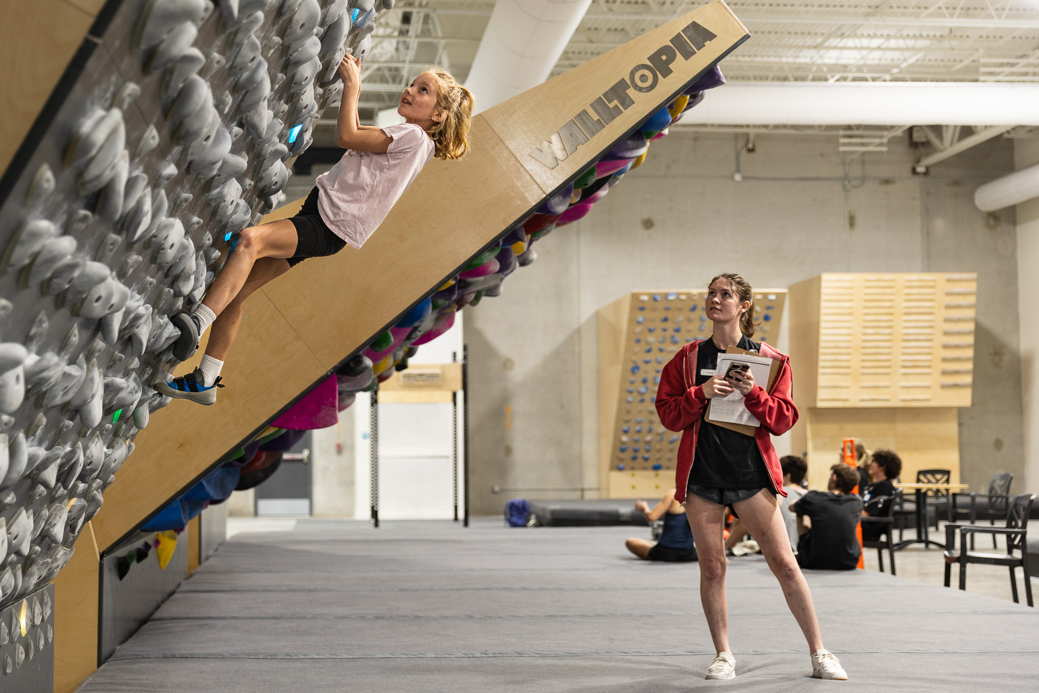 Young girl climbing on tilted wall, while female coach observes.